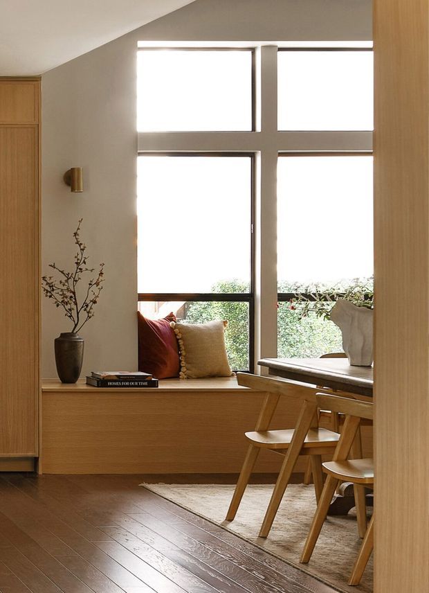 A warm, refined interior corner combining a built-in window seat, a compact dining area, and extensive timber joinery. Large black-framed cross-mullioned windows provide natural light, while the room features a timber dining table and chairs, a built-in timber bench with cushions, a neutral rug, a ceramic vase, and a small wall sconce. The palette is predominantly light and mid-tone wood, beige, taupe, and off-white, with a calm contemporary feel.