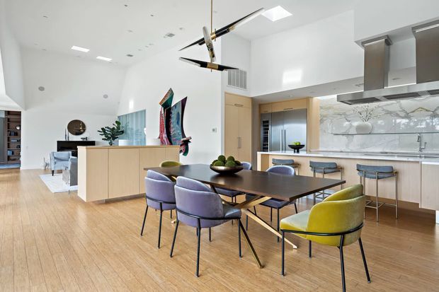 Bright open-plan kitchen and dining area with a large timber dining table, mixed upholstered dining chairs, pale timber flooring, and a central island. The contemporary white kitchen includes light timber cabinetry, a pale stone benchtop, a reflective stone or glass-look splashback, stainless-steel rangehood, and a long island with seating. A high white ceiling, skylights, modern pendant sculpture, and views into an adjacent living area create an airy, spacious interior.