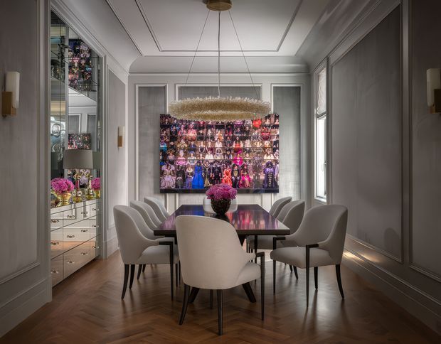 Elegant formal dining room with a rectangular dark dining table surrounded by upholstered sculptural dining chairs. The room features grey paneled walls, ornate ceiling moulding, a dark herringbone timber floor, tall windows, a large decorative chandelier, and a wall-mounted artwork or display above a console. Built-in glass-fronted cabinetry with drawers and decorative objects is visible along the left wall. The overall interior is refined, symmetrical, and richly styled.