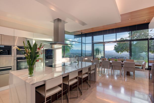 A spacious open-plan contemporary kitchen and dining area with a large pale stone island, integrated stainless-steel appliances, extensive floor-to-ceiling glazing, and a timber-lined ceiling section. The island includes multiple bar stools, while a separate dining table sits beside the windows. The polished floor and expansive glazing create a bright, refined interior with views toward a distant urban and mountainous landscape.