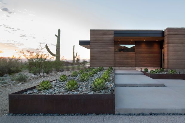 Modern single-storey house entrance at dusk, with a flat-roofed composition, dark horizontal timber-clad walls, recessed warm lighting, and a broad concrete path and steps leading to the front door. The foreground is a xeriscaped desert garden with agave and other succulents in gravel beds, steel-edged planters, and large cacti visible around the property.
