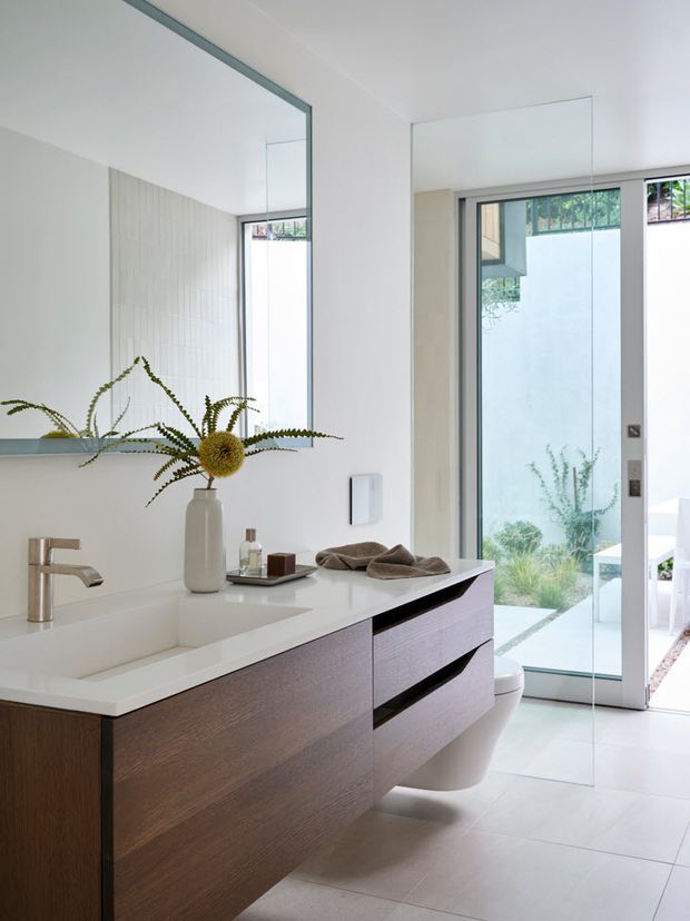 Bright contemporary bathroom with a floating dark timber vanity, white integrated countertop and basin, wall-mounted brushed metal tapware, large frameless mirror, pale tiled walls and floor, and a frameless glass shower enclosure. A glazed door opens to a landscaped outdoor area, adding natural light and an airy indoor-outdoor connection.