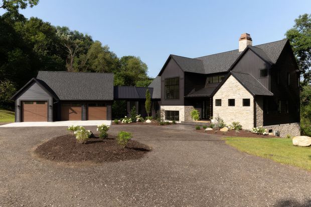 Exterior view of a large dark-clad residential home with steep gabled roofs, light stone masonry walls, a prominent chimney, and an attached detached-looking double garage connected by a covered link. The house sits within a landscaped, wooded setting with a broad gravel driveway and sparse new planting.