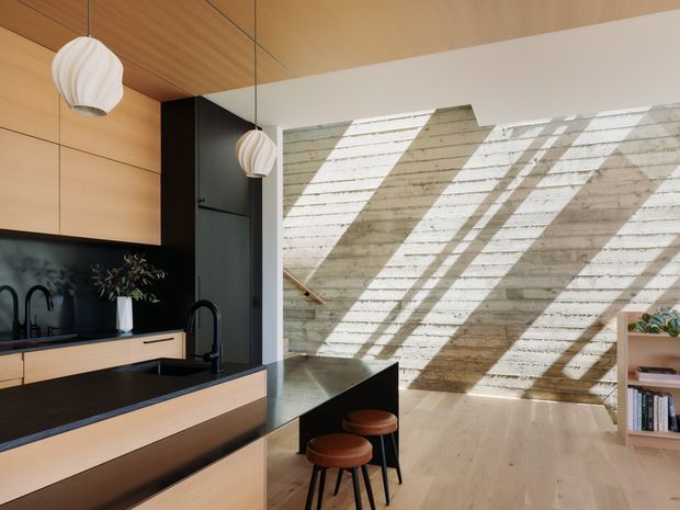 Warm, contemporary open-plan kitchen and living area with light timber cabinetry, a dark stone island and benchtops, black fixtures, pendant lights, and a dramatic exposed textured concrete feature wall illuminated by strong skylight-like shadows. A small dining or breakfast area with stools and shelving is visible beyond the kitchen.