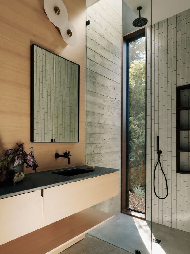 A contemporary bathroom with a floating timber vanity, matte black benchtop and black wall-mounted tapware. The walls combine warm timber panelling, pale vertically arranged rectangular tiles, and a textured concrete feature wall. A large black-framed mirror, sculptural wall lights, tall glazed window, open shower with black fixtures, and grey concrete floor complete the space. Natural greenery is visible outside.