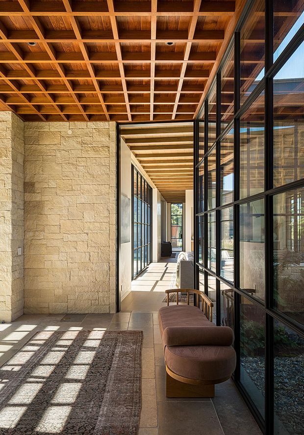 A long, sunlit interior corridor or living gallery with extensive black-framed glazing on the right, a textured pale stone feature wall on the left, and a warm timber coffered ceiling with exposed grid beams. The space includes a large patterned rug and a curved upholstered bench, with strong geometric shadows cast across the stone or tiled floor. The architecture has a refined indoor-outdoor connection and a warm, material-focused contemporary character.