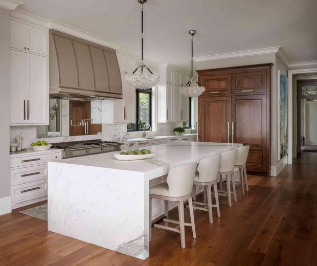 Elegant, spacious kitchen with white Shaker-style cabinetry, a large white marble-look island with seating for four, a professional range beneath a taupe vent hood, and crystal pendant lights. Warm dark timber flooring and a substantial traditional timber pantry or appliance cabinet create contrast, while a hallway is visible beyond the kitchen.