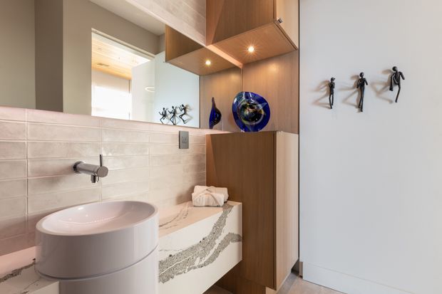 A refined contemporary bathroom vanity area with a round vessel basin on a white stone benchtop, wall-mounted tapware, pale textured rectangular wall tiles, and warm timber built-in cabinetry. Open display shelving contains decorative vessels, while a large mirror reflects the adjoining space. Sculptural black wall ornaments add a small accent to the otherwise neutral scheme.