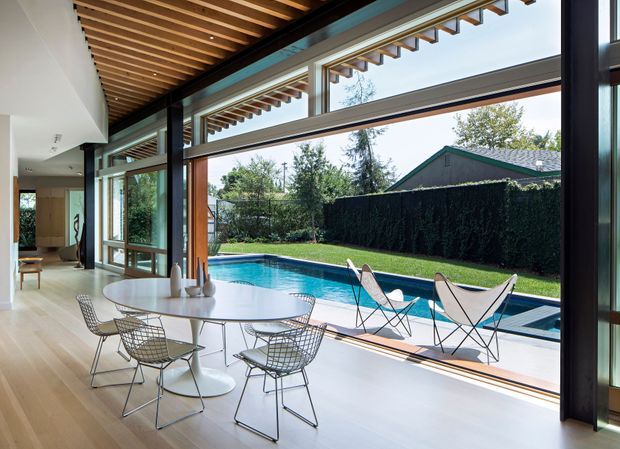 Bright, contemporary open-plan dining area with a round white dining table and wire-frame chairs beside expansive floor-to-ceiling glazing. The room opens directly to a landscaped backyard and swimming pool, with a timber slatted ceiling, pale timber flooring, dark structural window frames, and strong indoor-outdoor connection.
