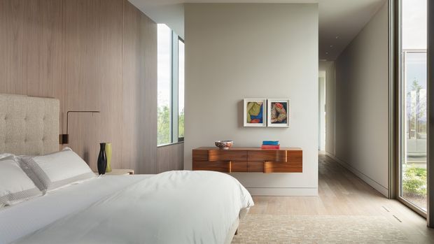 A bright, refined contemporary bedroom with a low-profile bed, upholstered headboard, pale bedding, full-height light timber wall panelling, and a floating mid-wood dresser. Large floor-to-ceiling windows and a glazed door bring in abundant natural light, while a narrow hallway or circulation space extends beyond the room. Minimal artwork and restrained furnishings create a calm, uncluttered interior.