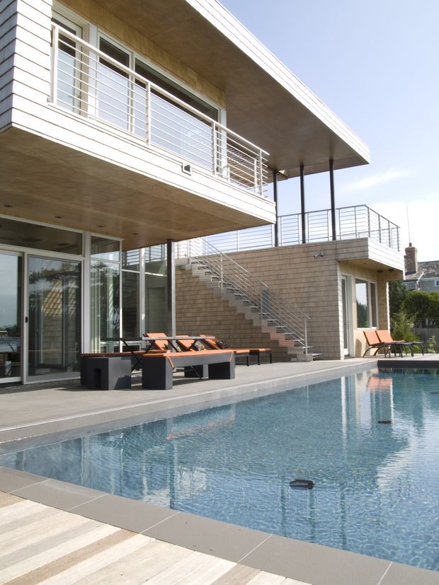 Exterior view of a modern multi-level residence centred on a swimming pool and elevated terrace. The building has broad projecting slab balconies, extensive glazing, light shingle-like wall cladding, pale timber soffits, slim dark supports, and horizontal metal balustrades. A stair rises from the pool deck to the upper terrace, with outdoor loungers arranged beside the pool.