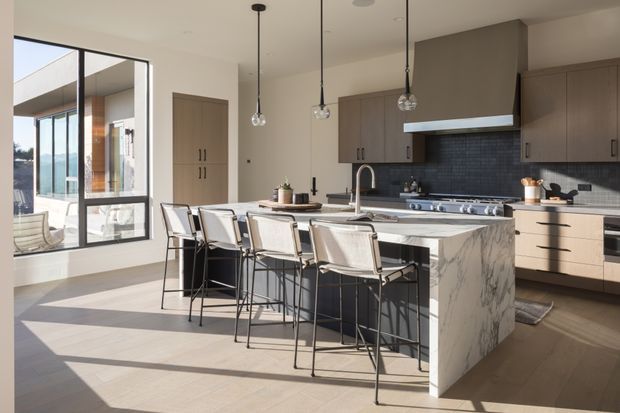 Bright contemporary kitchen with a large marble-look waterfall island, integrated dining-height seating for several people, taupe flat-panel cabinetry, dark tiled backsplash, stainless steel appliances, and pendant lights. Full-height glazing opens the room visually to an exterior terrace and water or landscape beyond.
