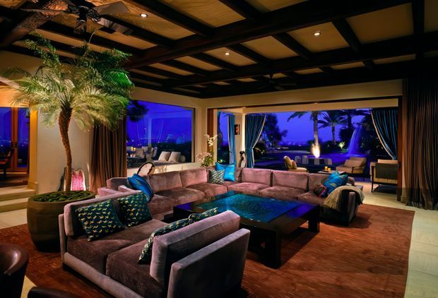 A luxurious, moody living room arranged around a large sectional sofa and low dark coffee table. The room features extensive glazing with views to a landscaped exterior, heavy curtains, indoor palms, warm timber ceiling panels set within a dark exposed-beam grid, recessed lighting, and rich purple-blue ambient illumination.
