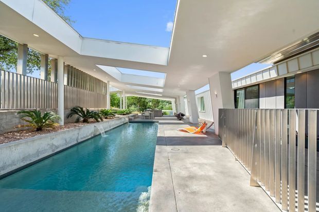 A bright contemporary residential outdoor area centered on a long narrow swimming pool, surrounded by white architectural roof planes and projecting covered walkways. Vertical metal privacy screens, concrete paving, large glazed openings, planted beds, and skylight-like roof openings create a structured indoor-outdoor setting. Lounge chairs and poolside furnishings are visible beneath the covered terrace.