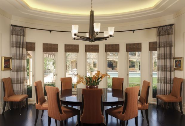 Elegant formal dining room with a large dark wood dining table surrounded by upholstered dining chairs. Tall bay-style windows with woven Roman blinds and plaid drapery overlook a pool and garden. The room features cream walls, dark timber flooring, framed artwork, and a prominent contemporary chandelier beneath a layered recessed ceiling with cove lighting.