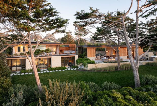 Wide exterior view of a low-slung contemporary residence integrated into a landscaped garden. The home features horizontal rooflines, extensive glazing, warm timber cladding, dark-framed windows, concrete or light masonry walls, and multiple outdoor terraces with built-in seating. Mature sculptural trees and layered planting frame the facade, while stepping-stone paths cross the lawn.