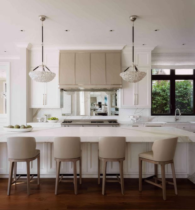 A bright, refined kitchen centered on a large white island with seating for four. The room features extensive white and warm greige built-in cabinetry, a mirrored splashback behind the range, dark timber flooring, black-framed windows, and two crystal pendant lights. Traditional architectural detailing such as decorative cornices, pilasters, and paneled cabinetry is blended with an elegant contemporary fit-out.