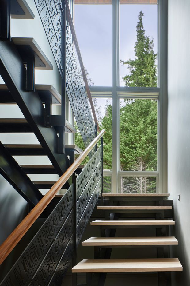 A contemporary interior stairway with open-riser timber treads supported by a dark steel structure. Decorative perforated or laser-cut dark metal balustrades and a warm timber handrail contrast with the light walls. Floor-to-ceiling glazing alongside the stairs provides abundant daylight and views of mature trees outside.