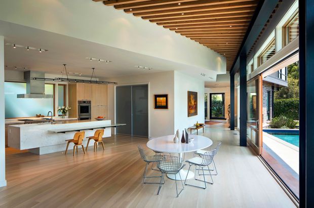 Open-plan contemporary kitchen, dining, and living area with a large stone island, timber cabinetry, round dining table, and wire-frame chairs. The room has extensive floor-to-ceiling glazing opening toward a swimming pool, a dramatic dark structural beam, and a warm timber slatted ceiling with integrated spotlights. A hallway and outdoor area are visible beyond the main space.