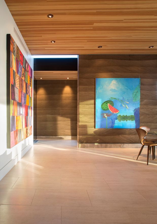 A sunlit contemporary interior hallway or open entry space with a wide pale wood or stone-look floor, a warm timber-lined ceiling with recessed spotlights, and a dark textured feature wall. Colourful artworks hang on the white side wall and the feature wall, while a small sculptural timber chair and table are partially visible at the right. Strong natural light enters from an unseen opening, creating a warm, gallery-like atmosphere.