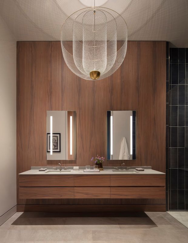 A refined contemporary bathroom vanity featuring a long double-sink floating timber joinery unit with a pale stone benchtop, twin illuminated rectangular mirrors, and a dramatic sculptural pendant light. The wall behind the vanity is clad in rich vertical-grain dark timber, while a narrow section of glossy dark rectangular tile is visible at right. The room has a warm, luxurious, symmetrical composition with neutral stone flooring and a curved, softly textured ceiling.