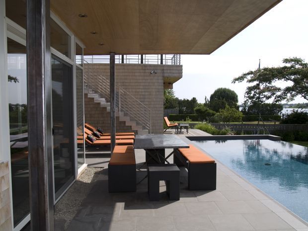 A contemporary outdoor pool terrace beside a modern house, viewed from under a broad timber-lined overhang. Floor-to-ceiling glazed doors open onto a paved entertaining area furnished with dark outdoor seating and orange cushions. A brick-clad stair tower with open metal balustrades leads to an upper level, while the rectangular swimming pool, clipped hedges, and landscaped garden extend toward a distant water view.