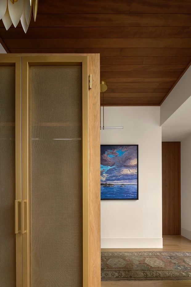 Interior view of a warm, refined hallway or entry area featuring a large built-in timber and mesh-panel door or cabinet in the foreground, a timber-lined ceiling, white plaster walls, timber flooring, a framed seascape artwork, and a patterned runner rug. A simple linear pendant light and dark timber doorway add architectural detail.