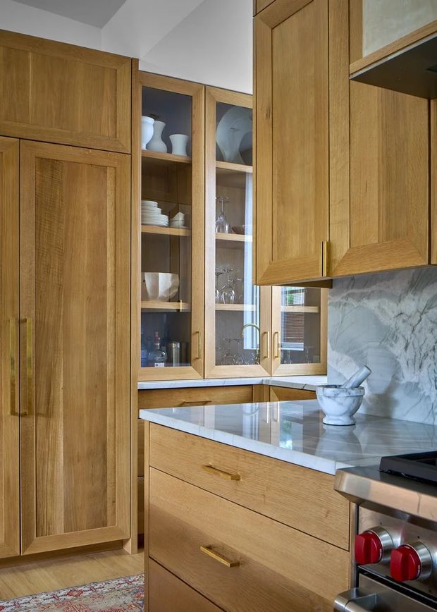 Close-up view of a warm timber kitchen with full-height oak cabinetry, integrated tall storage, glass-front display cabinets containing crockery and glassware, and brass-toned pulls. A pale veined stone benchtop and matching splashback frame the central preparation area, with a freestanding stainless-steel range partially visible at right. The room has a refined, traditional yet contemporary character and moderate natural light.