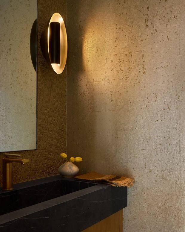 Close-up of a refined bathroom vanity featuring a dark stone benchtop and integrated basin, a warm brass/gold tap, and a sculptural illuminated wall sconce. The walls combine a textured dark feature panel with a pale, mottled plaster or concrete-like finish. Warm ambient lighting creates a luxurious, moody atmosphere, with a small decorative vase and folded textile on the counter.