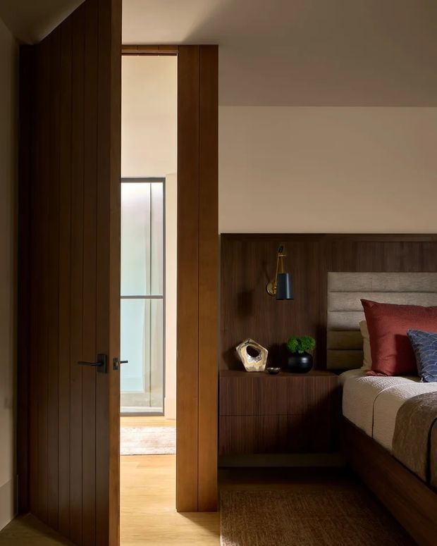 A warmly lit contemporary bedroom viewed through an open timber door. The room features dark vertical timber wall panelling and integrated bedside joinery, a built-in upholstered headboard, layered neutral and rust-toned bedding, a wall-mounted reading light, and a timber floor with a large rug. A bright adjoining space is visible through the doorway.