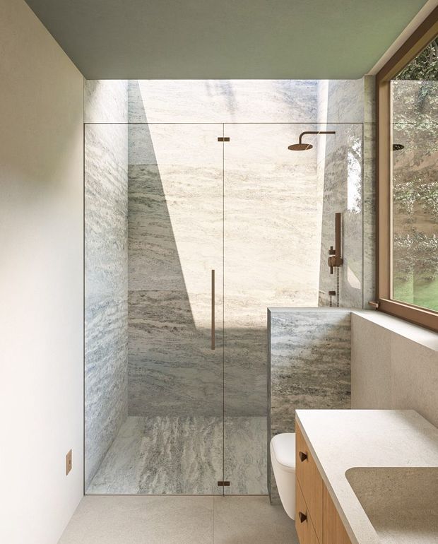 A compact contemporary bathroom featuring a walk-in glass shower clad in large-format grey and cream natural stone, with a rainfall shower and bronze-toned fittings. A timber vanity with a light stone benchtop sits beside the toilet, while a large timber-framed window brings in natural light and views of greenery.