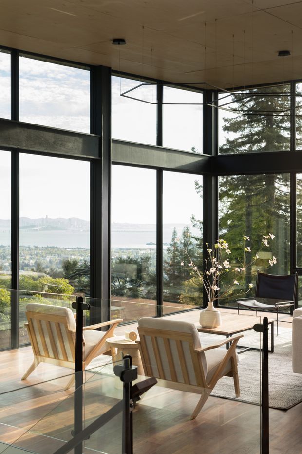 A bright, double-height living space with expansive black-framed floor-to-ceiling glazing overlooking a distant bay and landscape. Exposed dark structural beams divide the glazing, while timber ceiling panels, timber flooring, and mid-century style timber lounge chairs create a warm modern interior. A glass balustrade and stair or landing edge are visible in the foreground, along with a simple coffee table, vase of branches, and sculptural pendant lighting.