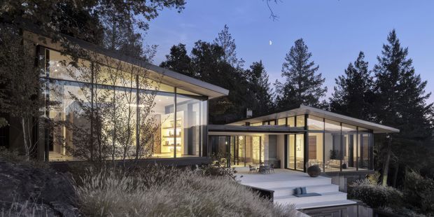 Contemporary single-storey residence set within a wooded landscape, composed of low-slung pavilion-like volumes with broad shallow-pitched roofs, extensive floor-to-ceiling glazing, and dark slender window frames. Warm interior lighting is visible through the glass, while a stepped terrace and landscaped planting connect the home to the surrounding trees.