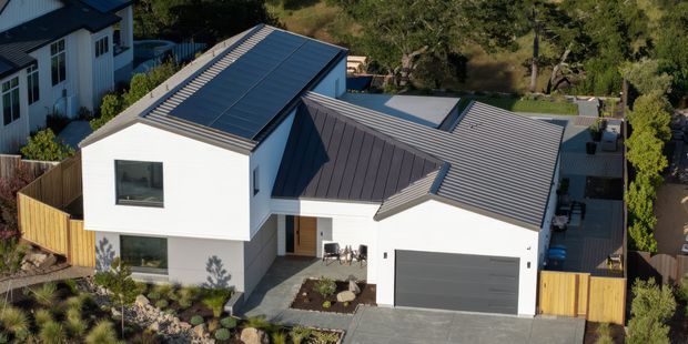 Aerial view of a contemporary multi-level residential home with crisp white rendered walls, dark standing-seam metal roof sections, and an extensive rooftop solar array. The home features angular roof forms, large rectangular windows, a recessed entry, an integrated single garage with a grey door, timber boundary fencing, paved driveway, and landscaped garden areas.