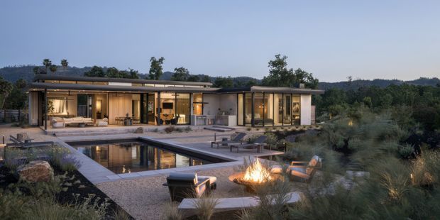 A low, single-storey contemporary residence set within a landscaped site, photographed at dusk. The home has extensive floor-to-ceiling glazing, a largely flat roofline, warm illuminated interiors, and a covered outdoor terrace opening onto a rectangular reflecting or swimming pool. Outdoor seating and a fire feature occupy the foreground, with dense natural planting surrounding the property.