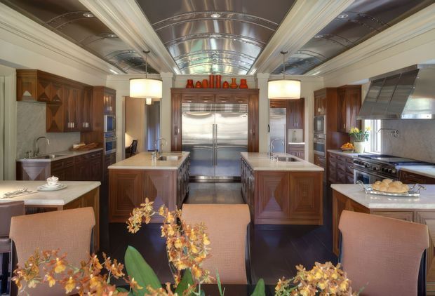 A richly detailed, symmetrical luxury kitchen with dark timber cabinetry, two central islands, integrated stainless-steel appliances, and a dramatic reflective metallic barrel-vault ceiling. Warm upholstered dining chairs and floral arrangements occupy the foreground, while decorative timber panelling, pendant lights, and polished surfaces create an ornate, refined atmosphere.
