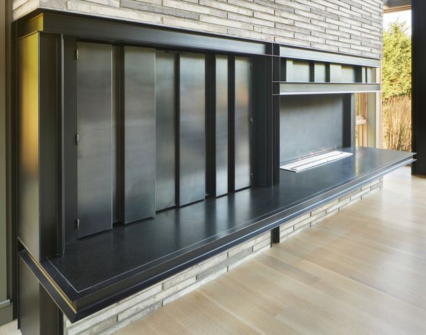 A contemporary built-in media and fireplace wall with dark vertical metal or timber-panelled joinery, a long dark stone-like hearth and shelf, and a linear recessed fireplace. The wall is faced in pale exposed brick, with warm light timber flooring and large glazing at the side. The composition is sleek, restrained, and strongly horizontal.