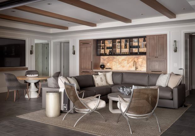 Sophisticated living room with a large charcoal sectional, woven lounge chairs, patterned area rug, and a small round coffee table. The room features white decorative wall panelling and cabinetry, dark timber ceiling beams, recessed lighting, and an integrated bar or kitchenette with a warm marble-look splashback, timber cabinetry, open display shelving, and brass-toned fittings. A dining nook and wall-mounted television are visible to the left.