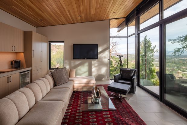 A warm, compact open-plan living room with a built-in kitchenette along the left wall, a large upholstered sectional sofa, black leather lounge chair, glass coffee table, wall-mounted television, and expansive floor-to-ceiling glazing. Timber-lined ceiling, dark-framed windows, neutral walls, and a richly patterned red rug create a refined modern interior with broad landscape views beyond the glass.