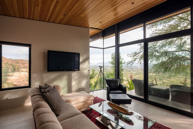 A warm, contemporary living room with a large sectional sofa, black leather lounge chair and glass coffee table. Floor-to-ceiling black-framed glazing provides expansive views of the surrounding landscape, while a timber-lined ceiling adds warmth above pale walls and large-format tiled flooring.