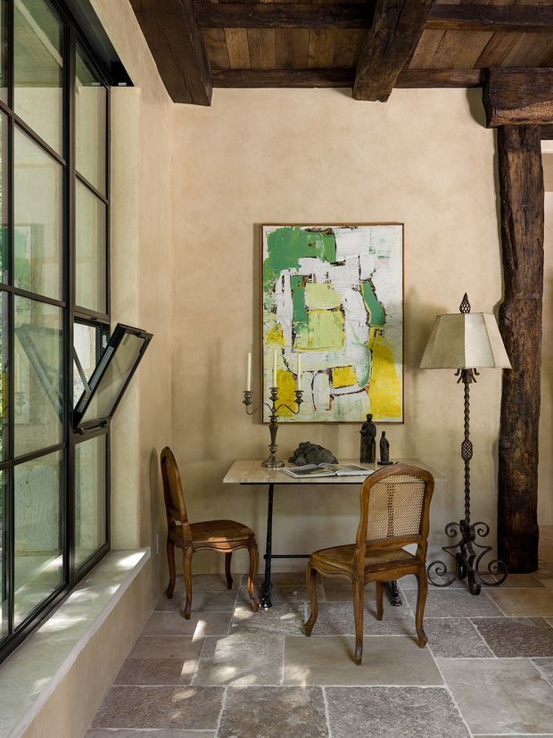 A refined rustic interior vignette with a small writing or dining table and two woven-back timber chairs set beside tall black-framed glazing. The room features thick exposed dark timber ceiling beams and a substantial timber post, warm textured plaster walls, large irregular stone floor tiles, an abstract artwork, and an ornate floor lamp. Natural light enters through the glazing, creating a warm, historic yet sophisticated atmosphere.