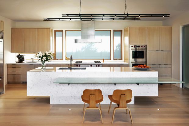 A bright, spacious contemporary kitchen with a large white stone island and integrated seating ledge, timber cabinetry, timber flooring, stainless-steel appliances, and a linear black suspended light fixture. Large windows bring in natural light, while a sculptural rangehood and waterfall-style island create strong focal points.
