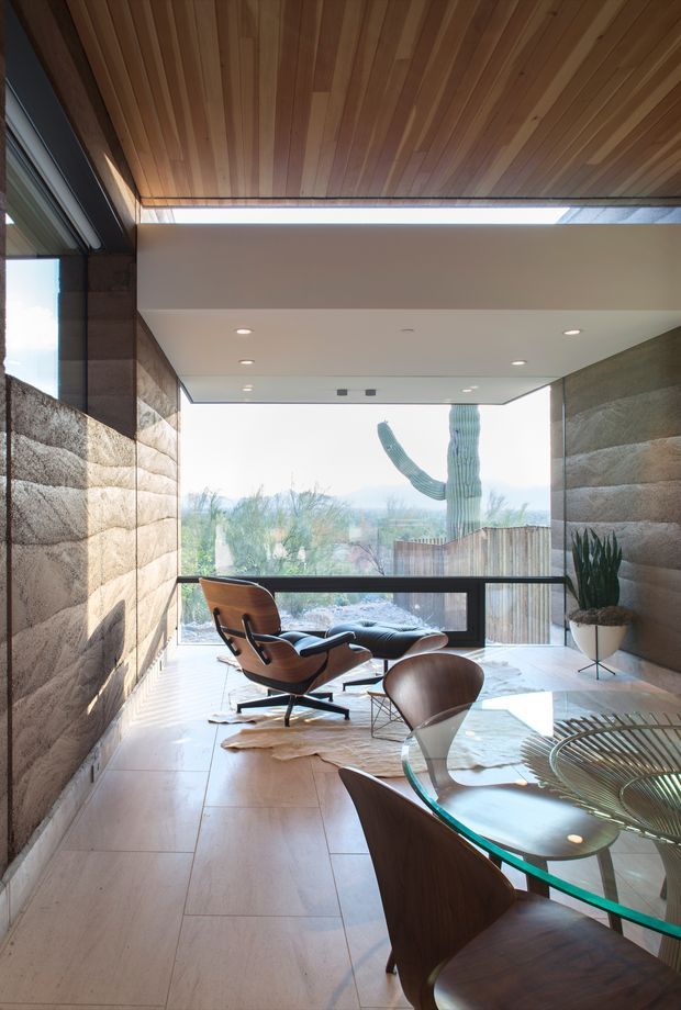 A refined modern interior combining a lounge and dining area. The space features floor-to-ceiling glazing overlooking a landscaped desert setting, a sculptural wooden ceiling with a concealed linear skylight, textured stone walls, large pale stone floor tiles, dark metal window framing, a mid-century-style lounge chair, and a round glass dining table with curved timber chairs.