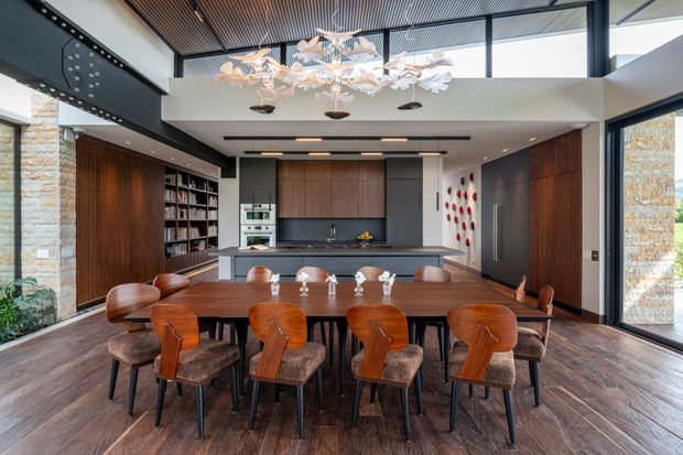 A sophisticated open-plan dining and kitchen interior with a large dark timber dining table surrounded by upholstered timber chairs. The kitchen features dark wood cabinetry, a grey island and benchtops, integrated appliances, and open shelving. Full-height glazing, exposed stone columns, a slatted timber ceiling, and a sculptural cluster pendant light create a warm, contemporary architectural setting.