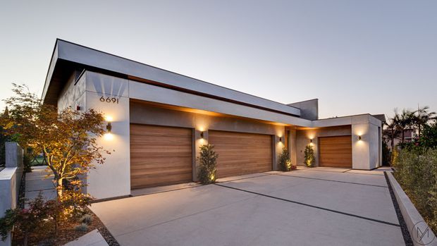 A dusk exterior view of a contemporary multi-car garage and residential frontage. The building has a long, low flat-roofed form, pale rendered walls, warm timber-toned sectional garage doors, recessed linear soffit lighting, and a broad concrete driveway. Minimal landscaping and integrated wall lights reinforce the modern architectural character.
