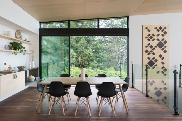 A bright contemporary dining area with a rectangular timber dining table and modern black chairs, positioned beside expansive black-framed glazing overlooking dense greenery. The room features dark timber flooring, a pale timber-lined ceiling, white walls, open shelving and light built-in cabinetry. A glass balustrade and geometric wall artwork are visible to the right.