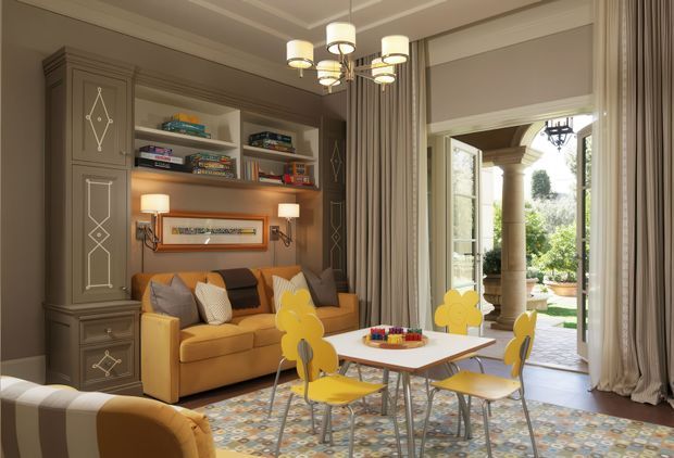 A refined family sitting and dining area with a mustard-yellow sofa, matching children's dining chairs, a white rectangular table, patterned area rug, and a bespoke dark taupe built-in shelving and cabinetry wall. Decorative wall panelling, framed artwork, layered curtains, and a contemporary multi-light chandelier create a polished, classic interior. Open French doors lead to a columned garden terrace.