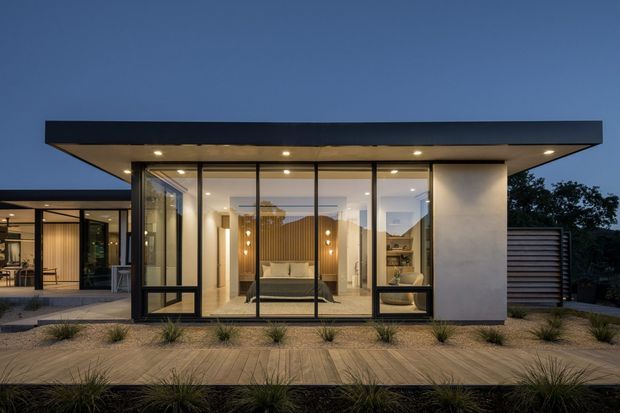 A contemporary single-storey pavilion-like house photographed at dusk, with a broad flat roof, extensive floor-to-ceiling glazing, dark metal frames, and pale rendered walls. The illuminated interior contains a living area with a sofa, timber feature wall and shelving. A covered outdoor area extends to the left, while a paved terrace and landscaped grasses occupy the foreground.