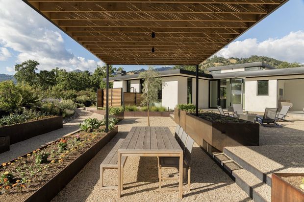 A contemporary single-storey residence viewed from a covered outdoor dining terrace. The deep timber-lined roof canopy frames a timber deck and dining table, with raised corten-toned garden beds, gravel paving, low planting, and stepped access to a light-rendered home. Black-framed glazing and a flat roof contribute to the modern, landscape-integrated composition, with wooded hills visible beyond.