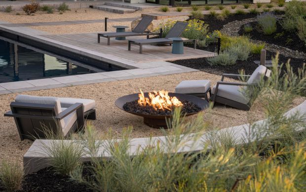 A landscaped outdoor pool area with a long rectangular swimming pool, timber-look decking, several contemporary sun loungers, and a circular fire pit seating area. Gravel paving, ornamental grasses, low shrubs, and dark planted beds create a structured, drought-tolerant landscape. The scene has a refined contemporary resort-like character with muted natural tones and warm firelight.